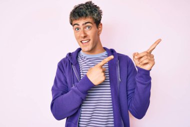 Genç, yakışıklı, sıradan, mor bir kazak giyen, gülümseyen ve kameraya bakıp iki elini ve parmağını yana doğru gösteren bir adam.. 