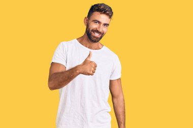 Latin asıllı genç bir adam, günlük beyaz tişört giyip mutlu bir el hareketi yapıyor. Kameraya bakıp başarıyı gösteren ifade. 