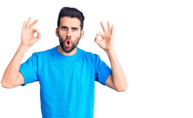 Sakallı, sıradan tişörtler giyen genç ve yakışıklı bir adam. Şaşırmış ve şaşırmış görünüyor. Parmaklarıyla onay sembolü yapıyor. çılgın ifade 