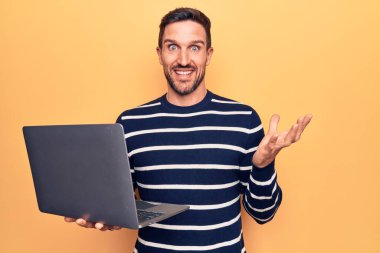 Genç yakışıklı adam dizüstü bilgisayarını kullanarak sarı arka planda duruyor başarıyı kutluyor mutlu bir gülümsemeyle ve kazanan yüz ifadesiyle elini kaldırarak.