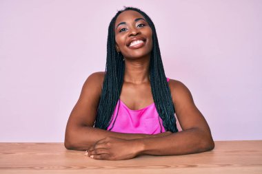 Saçları günlük kıyafetlerle örülü Afro-Amerikalı bir kadın masaya oturmuş, pozitif ve mutlu görünüyor. Kendine güvenen bir gülümsemeyle dişlerini gösteriyor. 