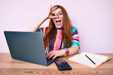 Ofiste çalışan genç kızıl saçlı kadın dizüstü bilgisayarla gülümsüyor mutlu mesut gülümsüyor. 