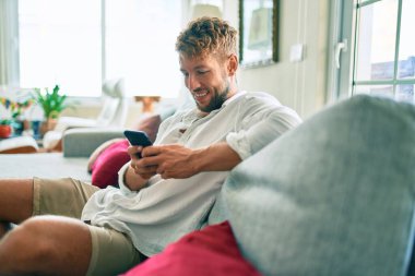 Yakışıklı beyaz adam mutlu gülümsüyor. Evdeki kanepede otururken akıllı telefon kullanıyor.