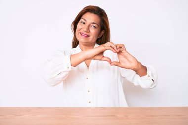 Orta yaşlı esmer, Latin iş kadını. Masada oturan, aşık bir şekilde gülümseyen, elleri ile kalp sembolü çizen sıradan beyaz tişörtlü kadın. romantik kavram. 