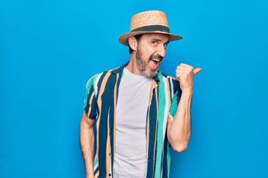 Orta yaşlı, yakışıklı, tatilde, gömlekli, şapkalı, mavi arka planda bir turist başparmağını yana kaldırmış, ağzı açık, mutlu bir şekilde gülümsüyor.