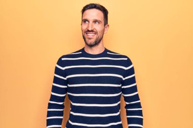 Sıradan çizgili süveter giyen genç yakışıklı adam, yalıtılmış sarı arka plan üzerinde duruyor, yan yana bakıyor, rahat profil pozu veriyor ve kendine güvenen bir gülümseme takınıyor..