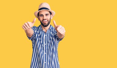 Sakallı, yaz şapkalı ve çizgili tişörtlü genç yakışıklı el hareketini onaylıyor, gülümsüyor ve başarı için mutlu. kazanan jest. 