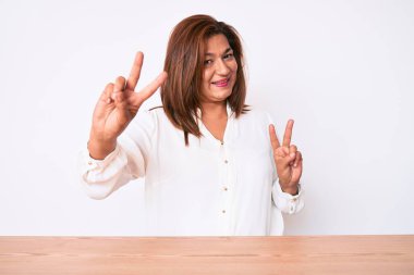 Orta yaşlı esmer, İspanyol iş kadını. Masada oturmuş gülümseyen, parmaklarını zafer işareti yaparken gösteren bir gülümseme takınıyor. İki numara.. 