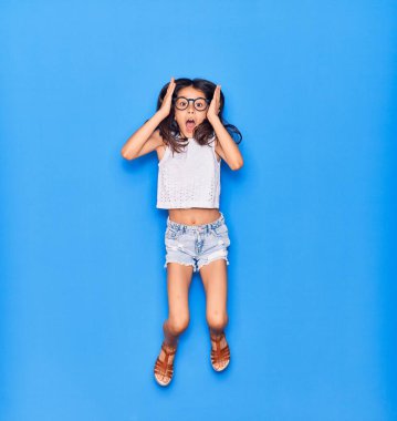 Günlük kıyafetler ve gözlük takan sevimli İspanyol kız ağzı açık kalınca şaşırdı. İzole edilmiş mavi arka planda eller havada zıplıyor.