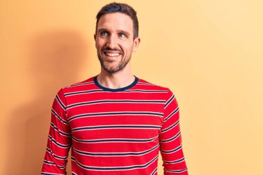 Sıradan çizgili tişört giyen genç yakışıklı adam izole edilmiş sarı arka planda dikilip yan yana bakıyor, rahat bir görünüm takınıyor ve kendine güvenen bir gülümseme sergiliyor..