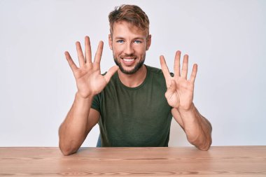 Gündelik giysiler giymiş genç beyaz bir adam masada oturuyor ve dokuz numaralı parmaklarını göstererek gülümsüyor. Kendinden emin ve mutlu.. 