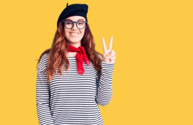 Fransız elbisesi giymiş genç bir kadın. Bere takmış, gülümseyen bir yüzle kameraya göz kırpıyor. Zafer işareti yapıyor. İki numara.. 