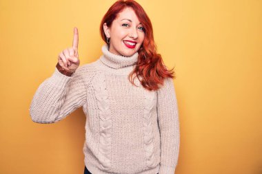Güzel, kızıl saçlı bir kadın sarı arka plan üzerine balıkçı yaka kazak giyiyor. Bir fikir veya soruyla gülümsüyor ve mutlu bir yüzle işaret ediyor.