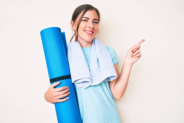 Güzel, beyaz bir kadın yoga minderi tutuyor. Gülümsüyor, mutlu bir şekilde elini ve parmağını yana doğru gösteriyor. 