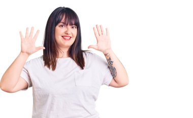 Genç ve cüsseli kadın gündelik kıyafetler giyer ve parmaklarını on numarayla gösterir ve gösterirken kendinden emin ve mutlu gülümser.. 