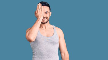 Mayolu ve kolsuz tişörtlü genç yakışıklı bir adam bir gözünü eliyle kapatıyor, yüzünde kendine güvenen bir gülümseme ve sürpriz bir duygu var.. 