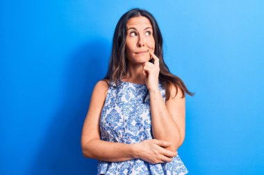 Genç, güzel, esmer bir kadın mavi arka plan üzerine gündelik kolsuz tişört giyip parmak çenesinde şüpheye odaklanmış ve kafasını kaldırıp,