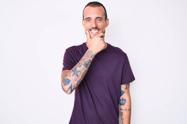 Dövmeli, sıradan kıyafetler giyen, kameraya kendinden emin, kolları çapraz ve çenesi havada gülümseyen genç yakışıklı bir adam. Olumlu düşünmek. 