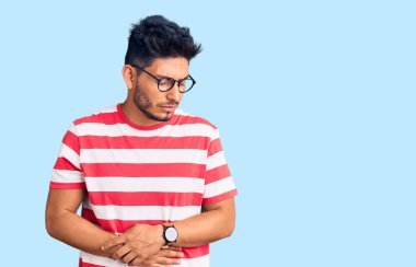 Yakışıklı Latin Amerikalı genç adam gündelik kıyafetler giyiyor ve elleri karnında gözlükler takıyor çünkü hazımsızlık, acı verici hastalıklar, kendini iyi hissetmiyor. Ağrı kavramı. 