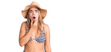 Bikini giyen ve şapkalı genç, sarışın bir kadın. İnançsızlıktan, şaşkınlıktan ve elleri çenesinde olan şaşkınlıktan büyülenmiş görünüyor. 