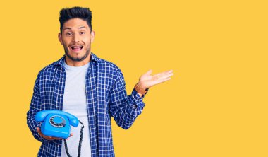 Yakışıklı Latin Amerikalı genç adam elinde klasik bir telefonla zaferi kutluyor mutlu bir gülümseme ve ellerini kaldırarak kazanan bir ifadeyle. 