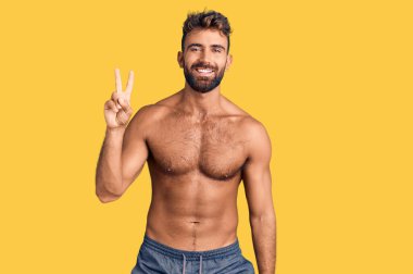 Genç İspanyol adam mayo giyiyor. İki numarayı gösterip işaret ediyor. Kendinden emin ve mutlu bir şekilde gülümsüyor.. 