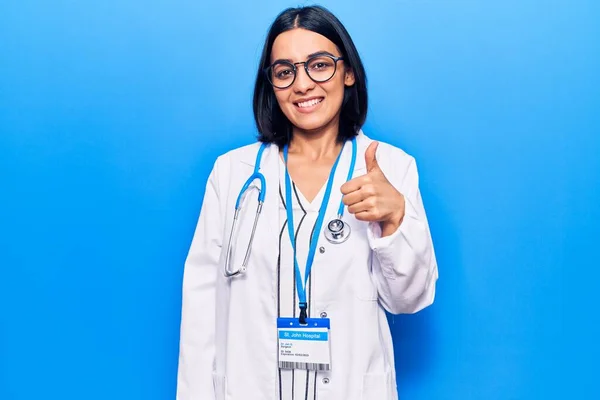 Young beautiful latin woman wearing doctor stethoscope and id card smiling happy and positive, thumb up doing excellent and approval sign