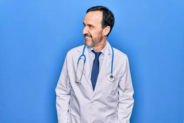 Middle age handsome doctor man wearing coat and stethoscope over isolated blue background looking to side, relax profile pose with natural face and confident smile.