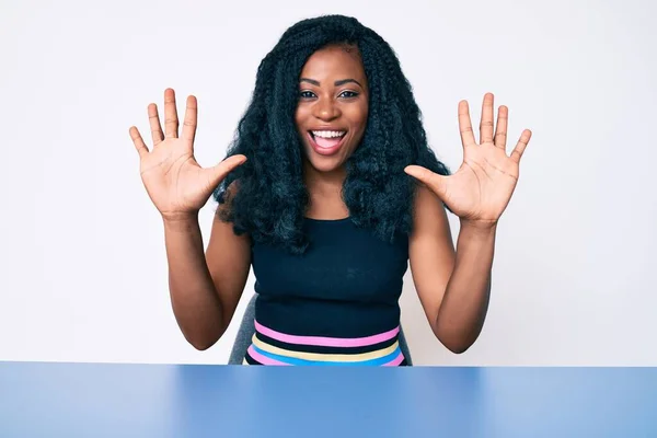 身穿休闲装的美丽的非洲女人坐在桌子上 用十号手指指着桌子 面带微笑 自信而快乐 — 图库照片