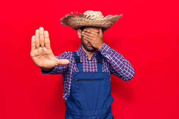 Young latin man wearing farmer hat and apron covering eyes with hands and doing stop gesture with sad and fear expression. embarrassed and negative concept.