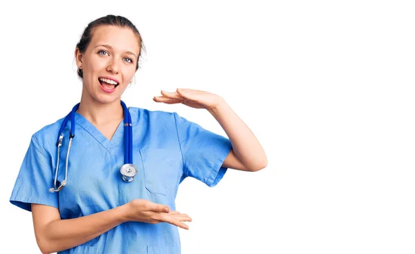 医師の制服を身に着けている若い美しいブロンドの女性と手でジェスチャーを聴診器は 大小のサインを示し シンボルを測定します カメラを見て笑ってる 概念を測定し — ストック写真