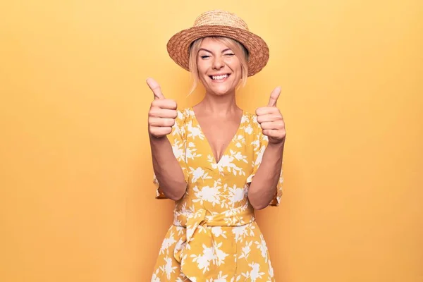 Beautiful blonde woman on vacation wearing summer hat and dress over yellow background success sign doing positive gesture with hand, thumbs up smiling and happy. Cheerful expression and winner gesture.
