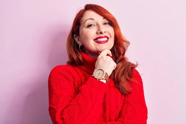 Young beautiful redhead woman wearing red casual turtleneck sweater over pink background smiling looking confident at the camera with crossed arms and hand on chin. Thinking positive.