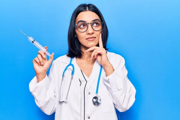 Young beautiful latin woman wearing doctor stethoscope holding syringe serious face thinking about question with hand on chin, thoughtful about confusing idea
