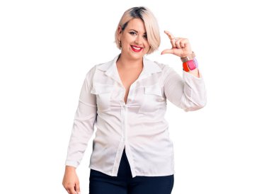 Genç, sarışın ve cüsseli bir kadın, rahat tişört giyiyor, gülümsüyor ve el hareketleriyle küçük beden işareti yapıyor, parmaklarına bakıyor ve kameraya bakıyor. ölçüm kavramı. 