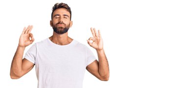 Gündelik beyaz tişört giyen genç İspanyol adam rahatla ve gözleri kapalı gülümse parmaklarıyla meditasyon hareketi yapıyor. yoga konsepti. 