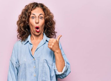 Orta yaşlı, güzel bir kadın pembe arka planda dikilen sıradan bir kot gömlek giyiyor. El parmağı yana dönük, ağzı açık, şaşkın bir ifade..