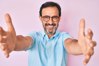 Orta yaşlı İspanyol bir adam günlük kıyafetler ve gözlüklerle kameraya bakıp kucaklaşmak için kollarını açmış gülümsüyor. Mutluluğu kucaklayan neşeli bir ifade.. 