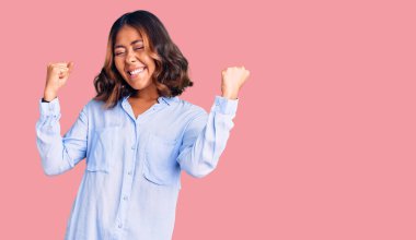 Genç, güzel, melez bir kadın günlük iş gömleği giyiyor. Çok mutlu ve heyecanlı. Kazanan bir jest yapıyor, kolları havada, gülümsüyor ve başarı için bağırıyor. kutlama konsepti. 