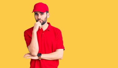 Sakallı, teslimat üniforması giyen, elleri çenesinde soru soran, düşünceli bir genç adam. Düşünceli bir yüzle gülümsüyor. şüphe kavramı. 