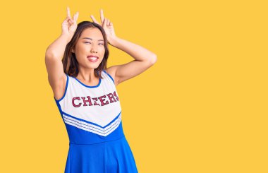 Genç, güzel Çinli kız amigo üniforması giyiyor. Komik ve çılgın pozlar veriyor. Parmakları kafasında tavşan kulağı gibi, neşeli gülüyor. 