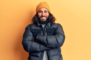 Gündelik kışlık giysiler giyen genç Arap adam kollarını kavuşturup kameraya bakarken gülümsüyor. pozitif kişi. 