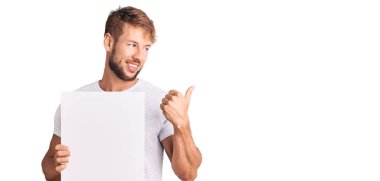 Kafkasyalı genç adam boş pankart tutuyor başparmağıyla yan tarafa bakıyor, ağzı açık gülümsüyor. 