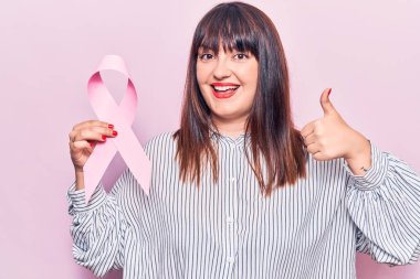 Elinde pembe kanser kurdelesi tutan genç ve cüsseli bir kadın mutlu ve pozitif gülümsüyor, başparmağı mükemmel ve onay işareti yapıyor. 