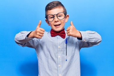 Sevimli sarışın çocuk inek papyonu ve gözlüklü başarı işareti takıyor. Elleriyle olumlu jest yapıyor. Gülümsüyor ve mutlu. Neşeli ifade ve kazanan jest. 