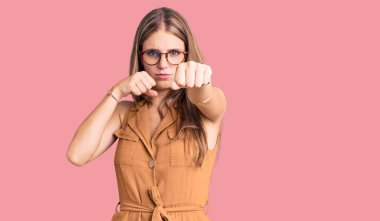 Genç, güzel, sarışın bir kadın. Günlük kıyafetler ve gözlük takıyor. Yumruk yumruğa dövüşüyor. 