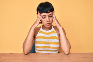 Masada oturan güzel esmer kadın sarı arka planda acı çektiği için çaresiz ve stresli baş ağrısından muzdarip. Eller başının üstünde.. 