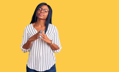 Genç Afro-Amerikalı kadın günlük kıyafetler ve gözlük takıyor ellerini birleştirip gülümseyerek gevşemiş ve neşeli gülümsüyor. Başarı ve iyimserlik 