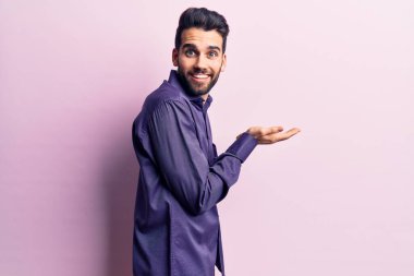 Sakallı, sıradan tişörtlü, elleri açık, fotokopi alanı gösteren ve mutlu bir şekilde gülümseyen genç yakışıklı bir adam. 