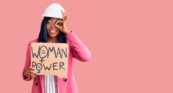 Young african american woman wearing architect helmet holding woman power banner smiling happy doing ok sign with hand on eye looking through fingers 